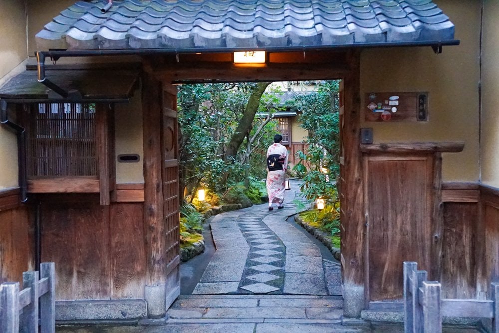 京都の町家の門越しに見える石畳の小径と歩く人の後ろ姿