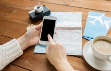 木の机で、地図と航空券の横に置いたスマートフォンを操作する手元