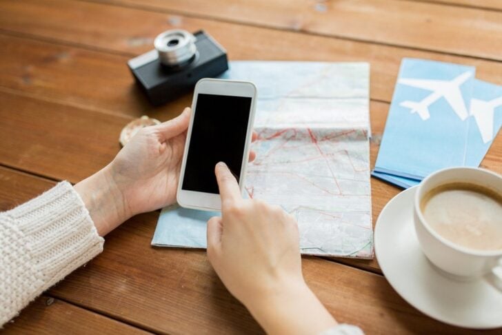 木の机で、地図と航空券の横に置いたスマートフォンを操作する手元