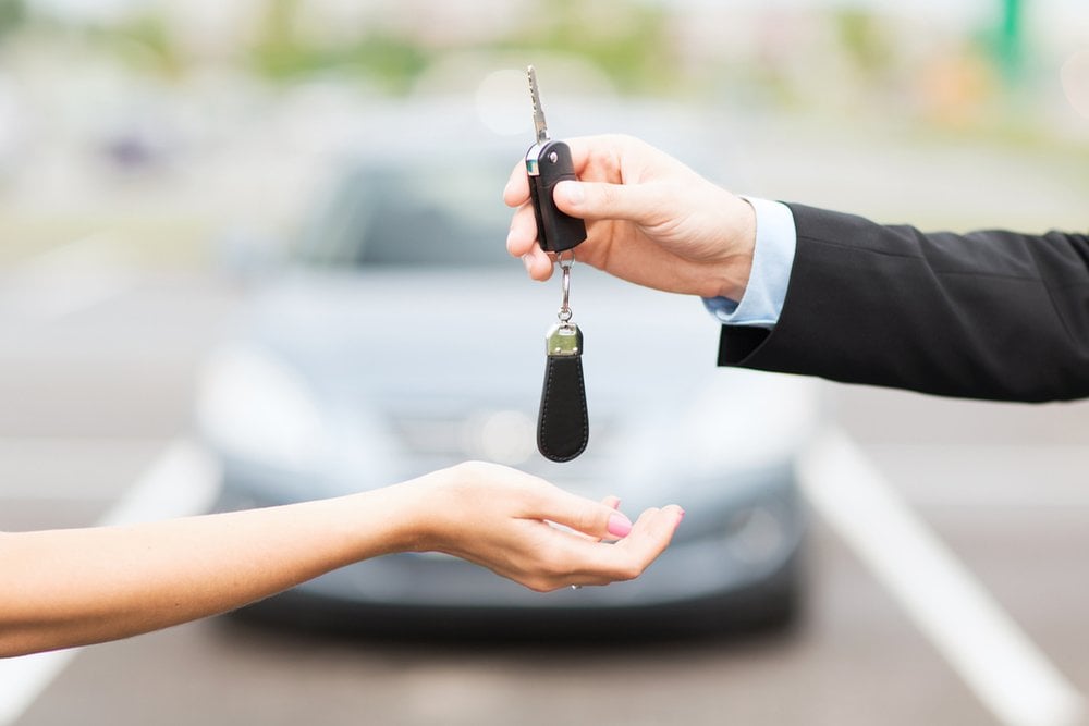 駐車場で車のキーを手渡す手元と、背景にぼやけた車