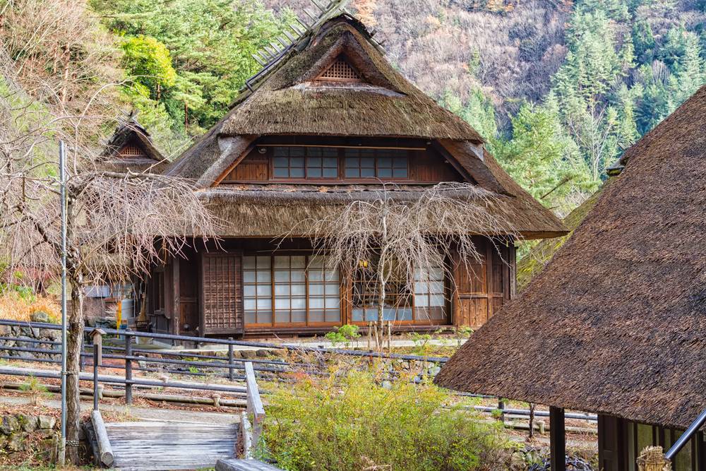 晩秋の山あいに建つ茅葺き屋根の古民家と木の小道